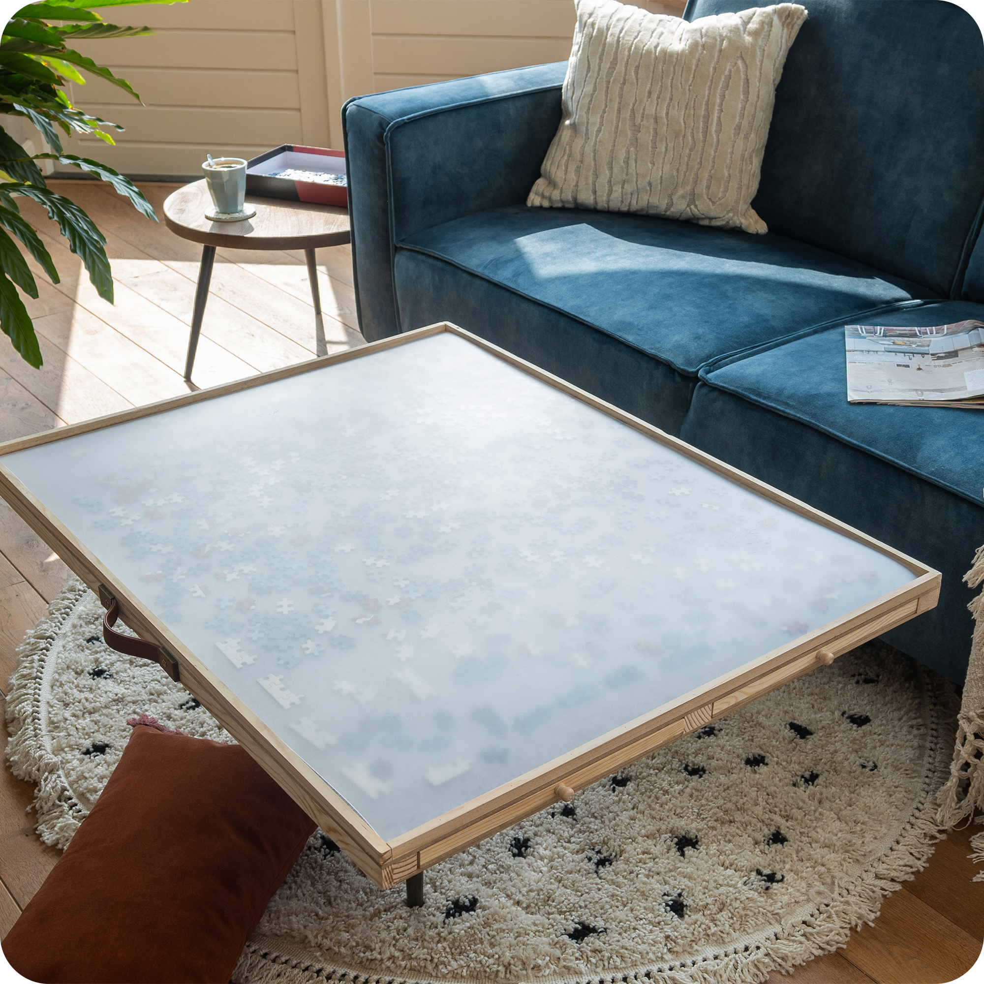 Een landelijke woonkamer met een blauwe bank en kussens. De verstelbare puzzelplaat staat op de salontafel met een doorzichtige beschermplaat en daaronder zitten de puzzelstukjes goed beschermd.